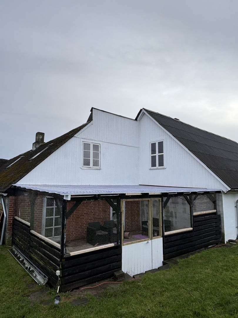 Overdækket træterrasse med sort træværk ved hvidt hus